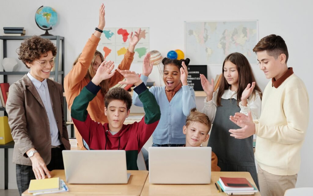 Grupo de estudiantes y una profesora en un aula, con dos niños usando laptops y el resto celebrando con las manos en alto, mostrando alegría en un entorno de aprendizaje tecnológico