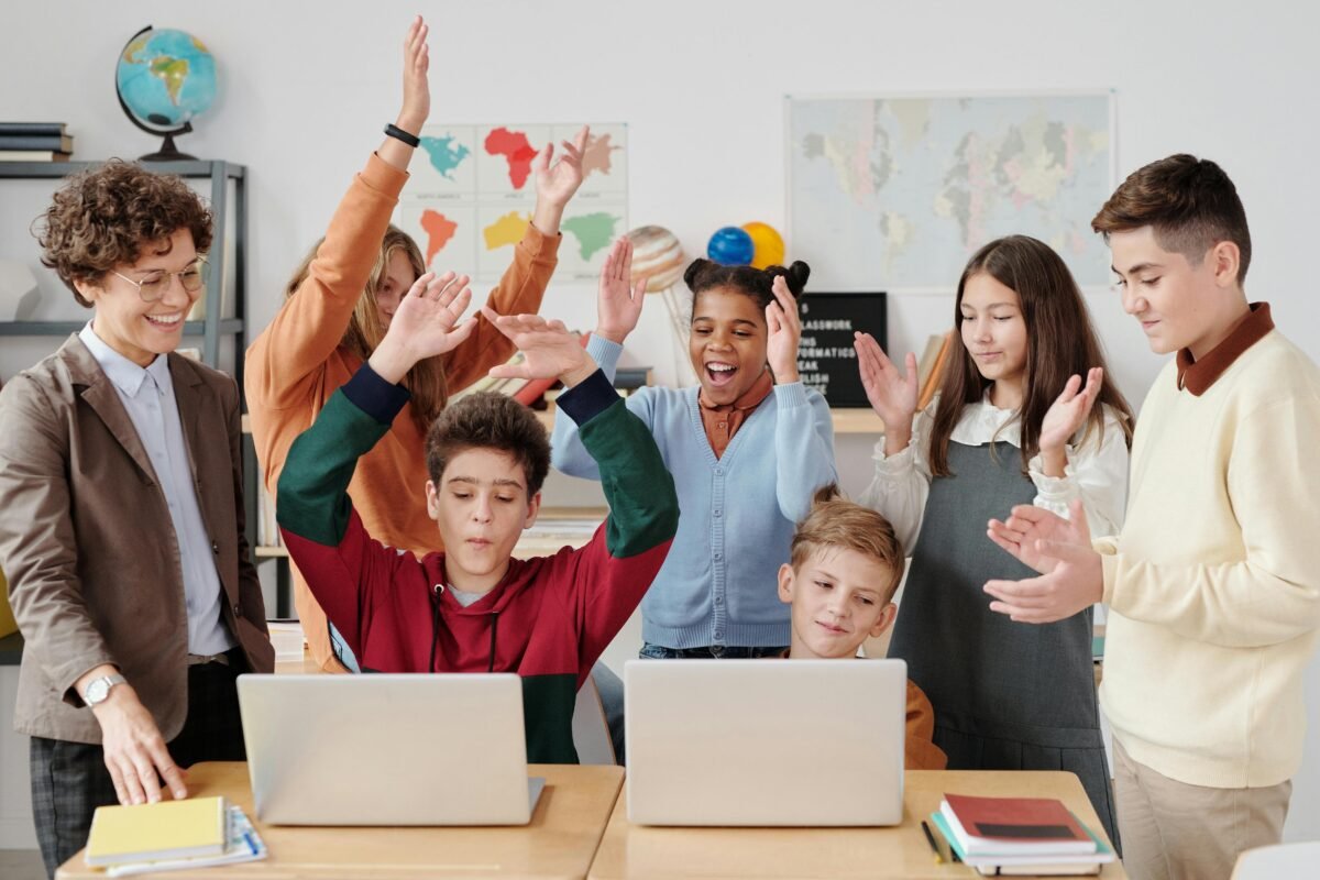 Grupo de estudiantes y una profesora en un aula, con dos niños usando laptops y el resto celebrando con las manos en alto, mostrando alegría en un entorno de aprendizaje tecnológico