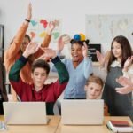 Grupo de estudiantes y una profesora en un aula, con dos niños usando laptops y el resto celebrando con las manos en alto, mostrando alegría en un entorno de aprendizaje tecnológico