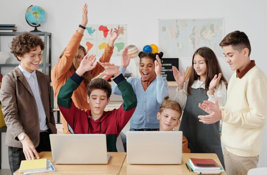Grupo de estudiantes y una profesora en un aula, con dos niños usando laptops y el resto celebrando con las manos en alto, mostrando alegría en un entorno de aprendizaje tecnológico
