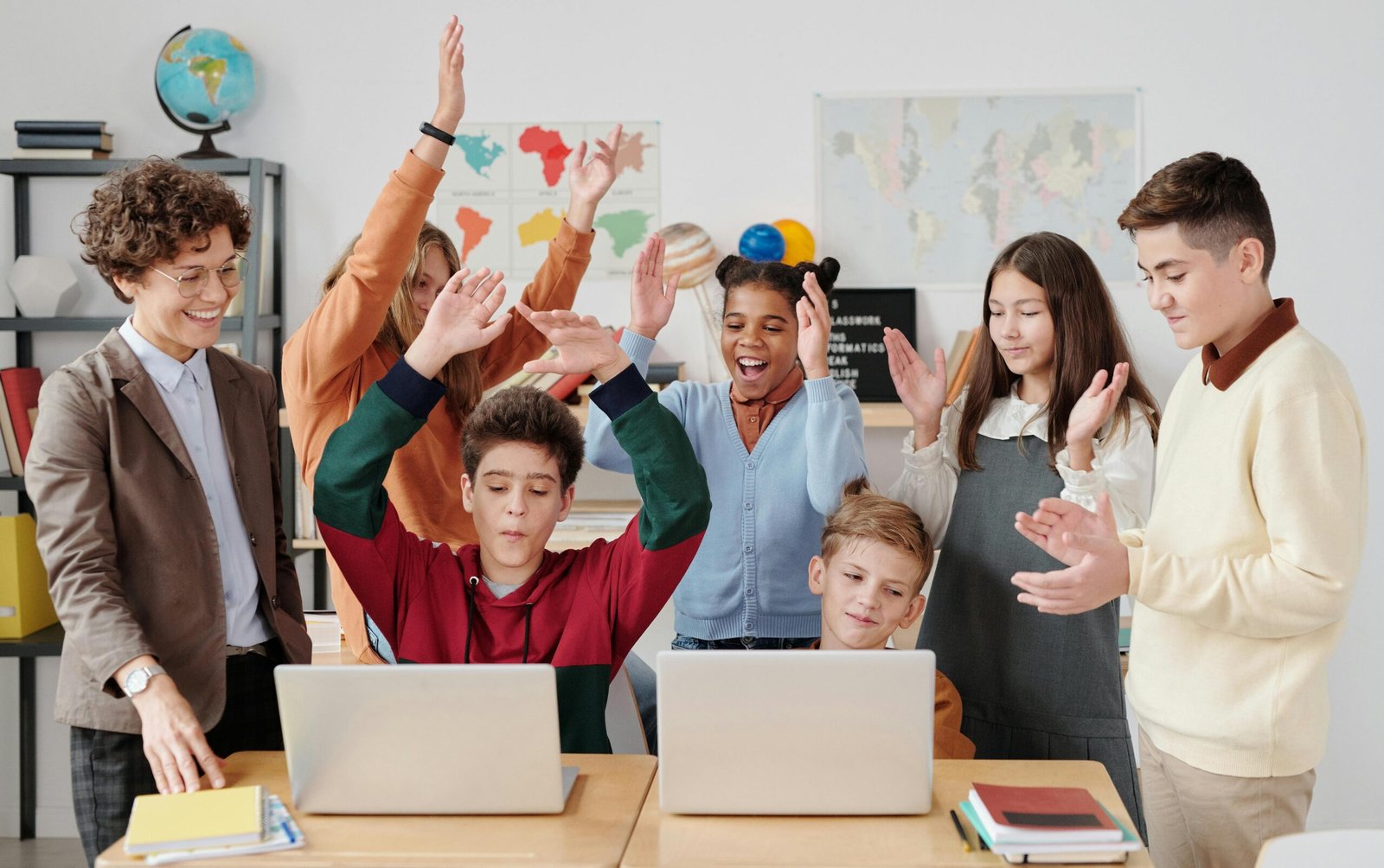 Grupo de estudiantes y una profesora en un aula, con dos niños usando laptops y el resto celebrando con las manos en alto, mostrando alegría en un entorno de aprendizaje tecnológico