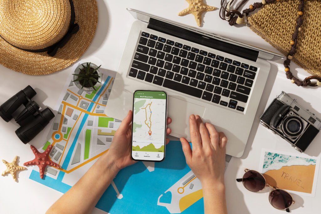 Vista superior de una persona sosteniendo un smartphone con una aplicación de mapas, mientras planifica un viaje con una laptop, un mapa de papel, binoculares y una cámara sobre una mesa.