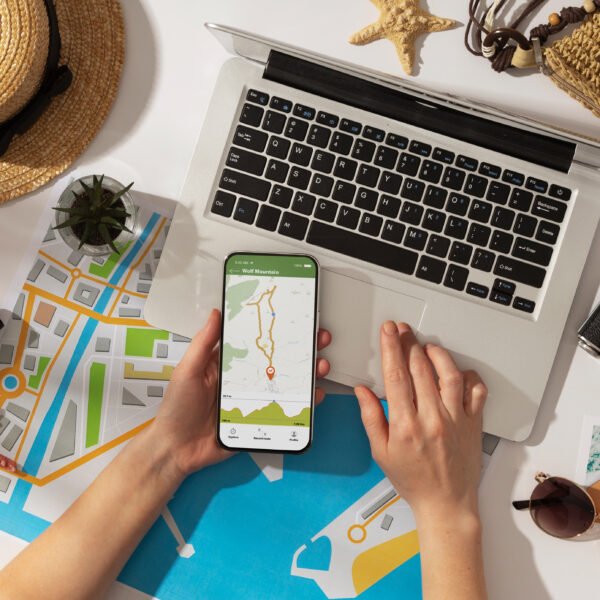 Vista superior de una persona sosteniendo un smartphone con una aplicación de mapas, mientras planifica un viaje con una laptop, un mapa de papel, binoculares y una cámara sobre una mesa.