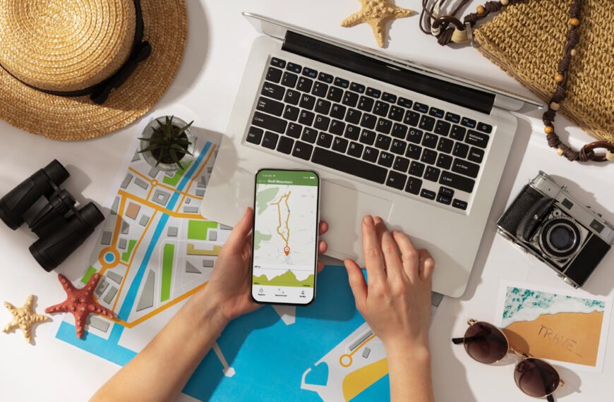 Vista superior de una persona sosteniendo un smartphone con una aplicación de mapas, mientras planifica un viaje con una laptop, un mapa de papel, binoculares y una cámara sobre una mesa.