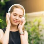 Mujer sonriente con audífonos blancos disfrutando de música con cancelación de ruido en un parque soleado.
