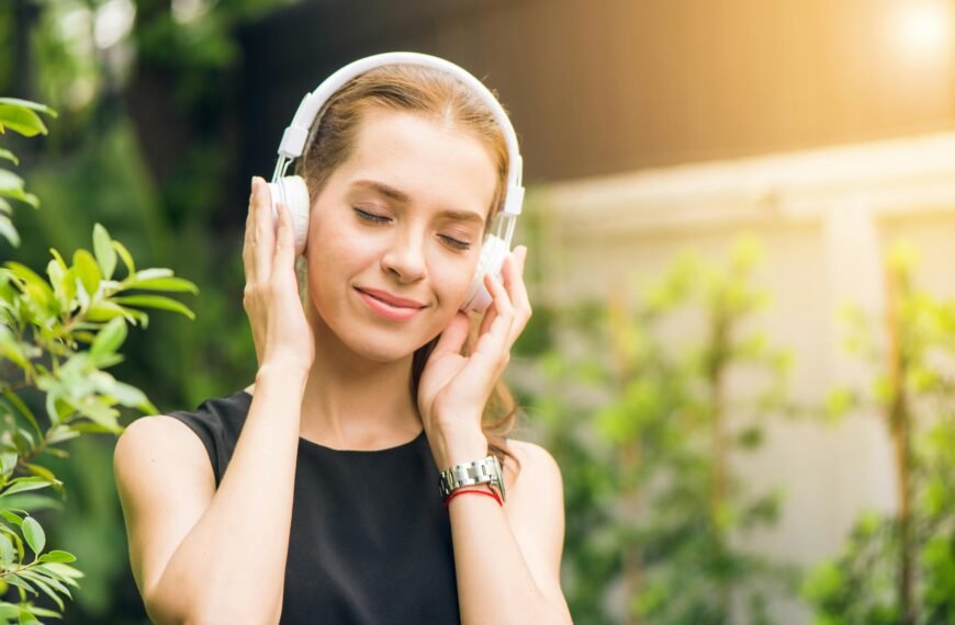 Mujer sonriente con audífonos blancos disfrutando de música con cancelación de ruido en un parque soleado.